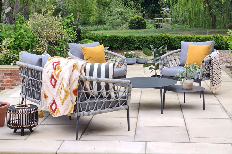 Luxury rope garden furniture (two armchairs, a sofa and two coffee tables) with mustard yellow cushions sit outside on a patio.